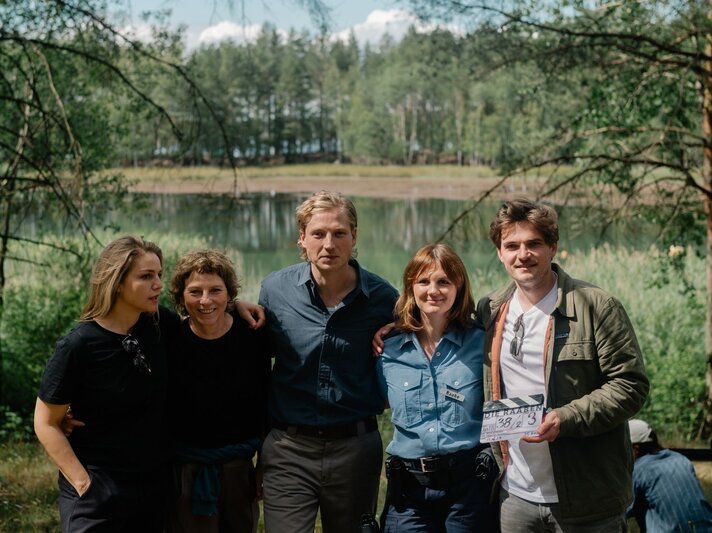 Fünfköpfiges Filmteam steht lachend vor einem idyllischen See, eine Frau in Polizeiuniform trägt ein Namensschild mit „Raabe“. | © rbb/Elliott Kreyenberg