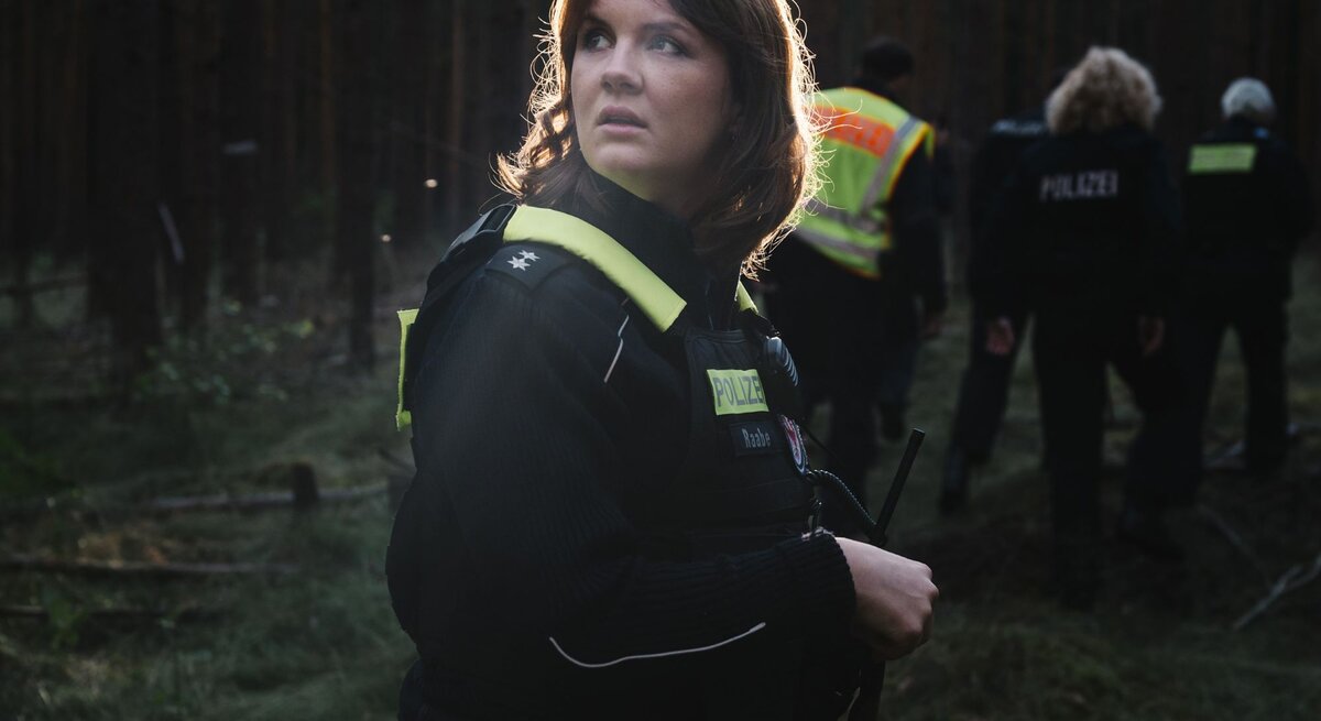 In der Abenddämmerung durchkämmt ein Polizeiteam einen dichten Wald, eine Frau im Vordergrund hält ein Funkgerät und wirkt alarmiert.