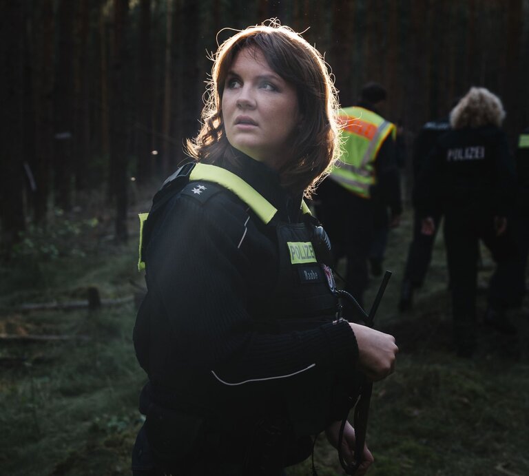 In der Abenddämmerung durchkämmt ein Polizeiteam einen dichten Wald, eine Frau im Vordergrund hält ein Funkgerät und wirkt alarmiert. | © rbb/Elliott Kreyenberg