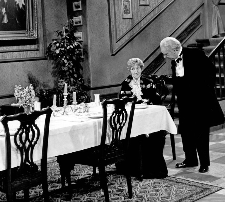 Ein älterer Mann im Anzug beugt sich zu einer elegant gekleideten Dame an einem festlich gedeckten Tisch in einem klassischen Speisesaal. | © IMAGO / United Archives