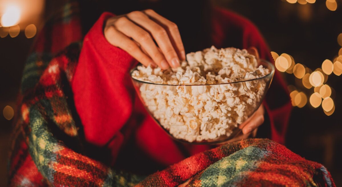 Eine Person in rotem Pullover und karierter Decke greift in eine Schüssel Popcorn vor unscharfen, warm leuchtenden Lichterketten, während sie einen Weihnachtsfilm schaut.