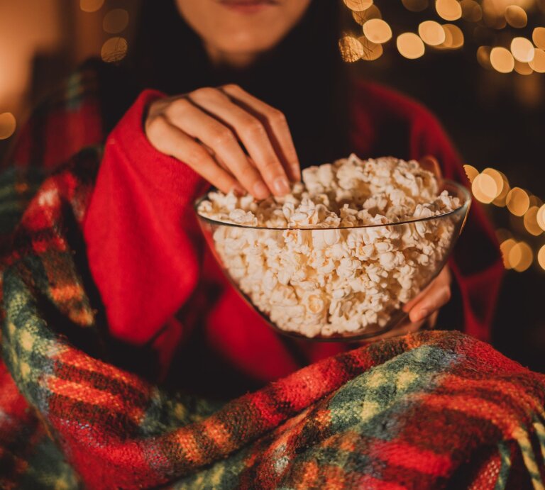 Eine Person in rotem Pullover und karierter Decke greift in eine Schüssel Popcorn vor unscharfen, warm leuchtenden Lichterketten, während sie einen Weihnachtsfilm schaut. | © Adobe Stock/ Andrii Lysenko