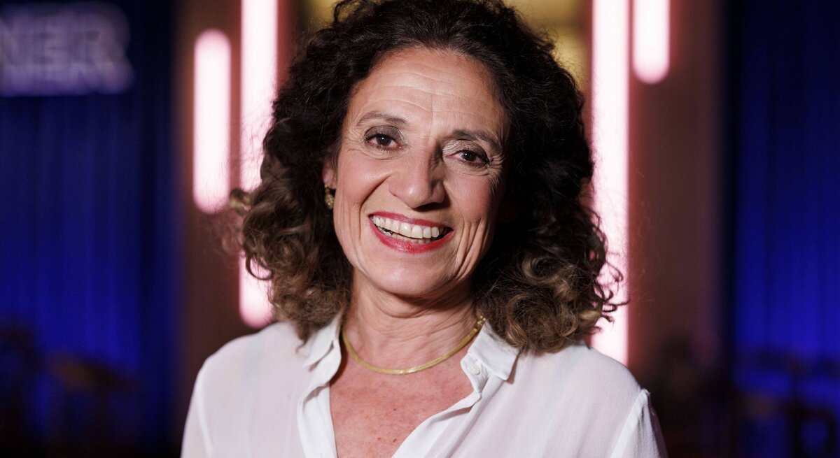  Das Bild zeigt Adriana Altaras, eine lächelnde Frau mit lockigem dunklem Haar in einem weißen Hemd. Im Hintergrund sind unscharfe Lichter und ein leicht erkennbarer Schriftzug zu sehen, was darauf hindeutet, dass die Aufnahme bei einem Event oder in einer Umgebung mit Bühnenbeleuchtung gemacht wurde. Die Frau strahlt mit einem breiten Lächeln, und die Beleuchtung hebt ihre freundlichen Gesichtszüge hervor.