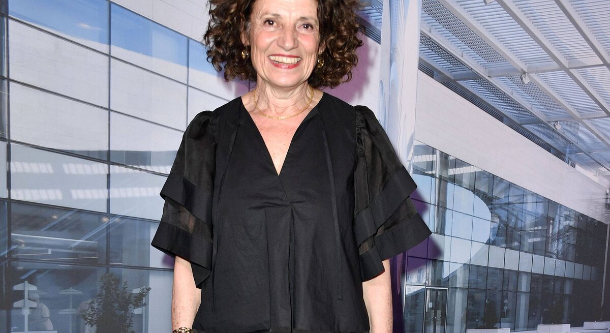 Adriana Altaras hat lockiges dunkles Haar und trägt ein schwarzes Outfit. Sie steht vor dem Hintergrund eines modernen Gebäudes mit Glasfassade und Metallstrukturen. Sie trägt ein stilvolles, luftiges Oberteil mit Rüschenärmeln und ein Lächeln, das Wärme und Begeisterung ausstrahlt. Sie trägt auch ein Armband und eine Halskette, was ihren eleganten Look vervollständigt.