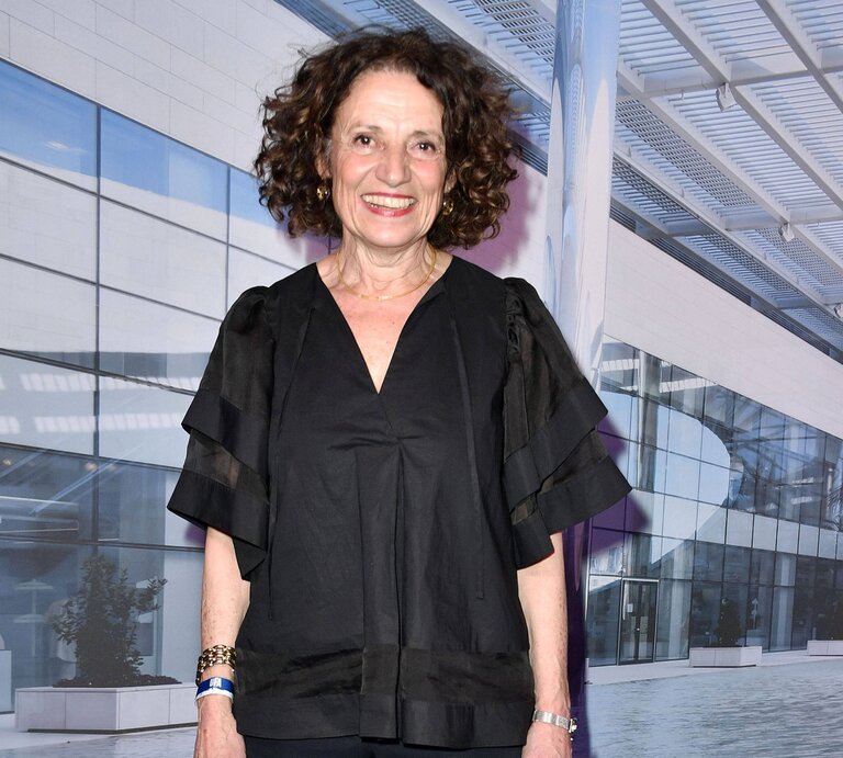 Adriana Altaras hat lockiges dunkles Haar und trägt ein schwarzes Outfit. Sie steht vor dem Hintergrund eines modernen Gebäudes mit Glasfassade und Metallstrukturen. Sie trägt ein stilvolles, luftiges Oberteil mit Rüschenärmeln und ein Lächeln, das Wärme und Begeisterung ausstrahlt. Sie trägt auch ein Armband und eine Halskette, was ihren eleganten Look vervollständigt. | © IMAGO / Future Image