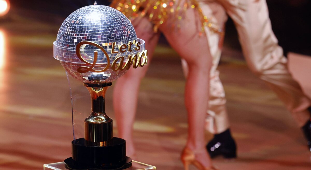 Ein glitzernder Disco-Ball-Trophäe auf einem Podest mit der Aufschrift "Let's Dance", im Hintergrund unscharf die Beine zweier Tänzer in Bewegung, einer trägt schwarze Schuhe und der andere goldene Tanzschuhe mit Glitzer.