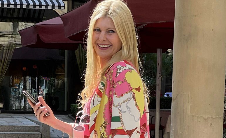 Das Bild zeigt eine Frau mit langen blonden Haaren und einem strahlenden Lächeln, die in einer städtischen Umgebung steht. Sie trägt ein farbenfrohes Outfit mit einem Muster, das an Ketten und Riemen erinnert, und strahlt eine fröhliche und entspannte Stimmung aus. | © privat