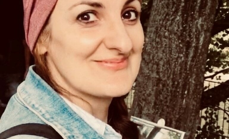 Die Frau auf dem Foto trägt ein rosa Kopftuch und eine Jeansjacke. Sie hat dunkles, zur Seite geflochtenes Haar und ein freundliches Lächeln. Der Hintergrund ist unscharf, aber es scheint, als befände sie sich im Freien, möglicherweise in einem Park, da Bäume zu sehen sind.  | © privat