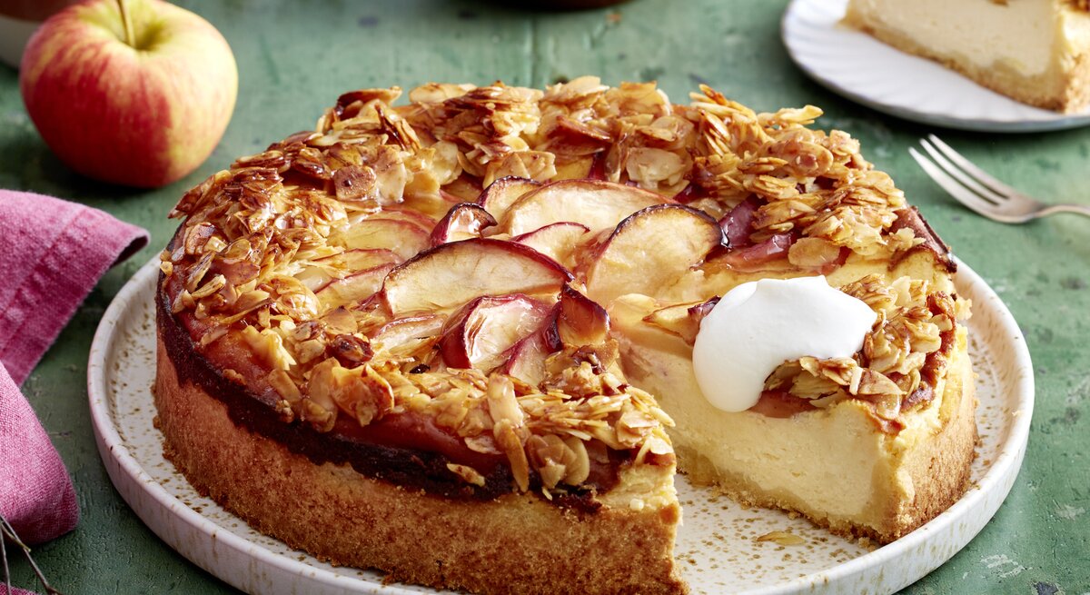 Apfel-Quark-Bienenstich auf einem weißen Teller auf grünem Hintergrund. Links ein pinkes Geschirrtuch und ein Apfel.