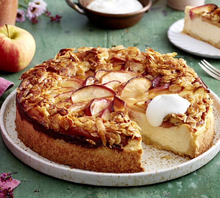 Apfel-Quark-Bienenstich auf einem weißen Teller auf grünem Hintergrund. Links ein pinkes Geschirrtuch und ein Apfel. | © EAT CLUB/Isabell Triemer
