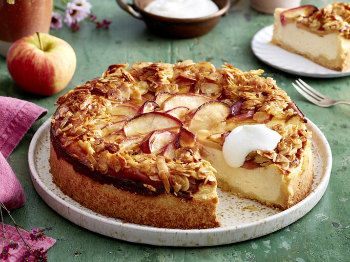 Apfel-Quark-Bienenstich auf einem weißen Teller auf grünem Hintergrund. Links ein pinkes Geschirrtuch und ein Apfel. | © EAT CLUB/Isabell Triemer