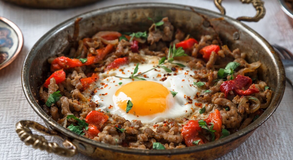 Kiymali Yemek mit Spiegelei und frischen Kräutern in einer runden Pfanne aus Metall / Gabel / Untergrund Holz
