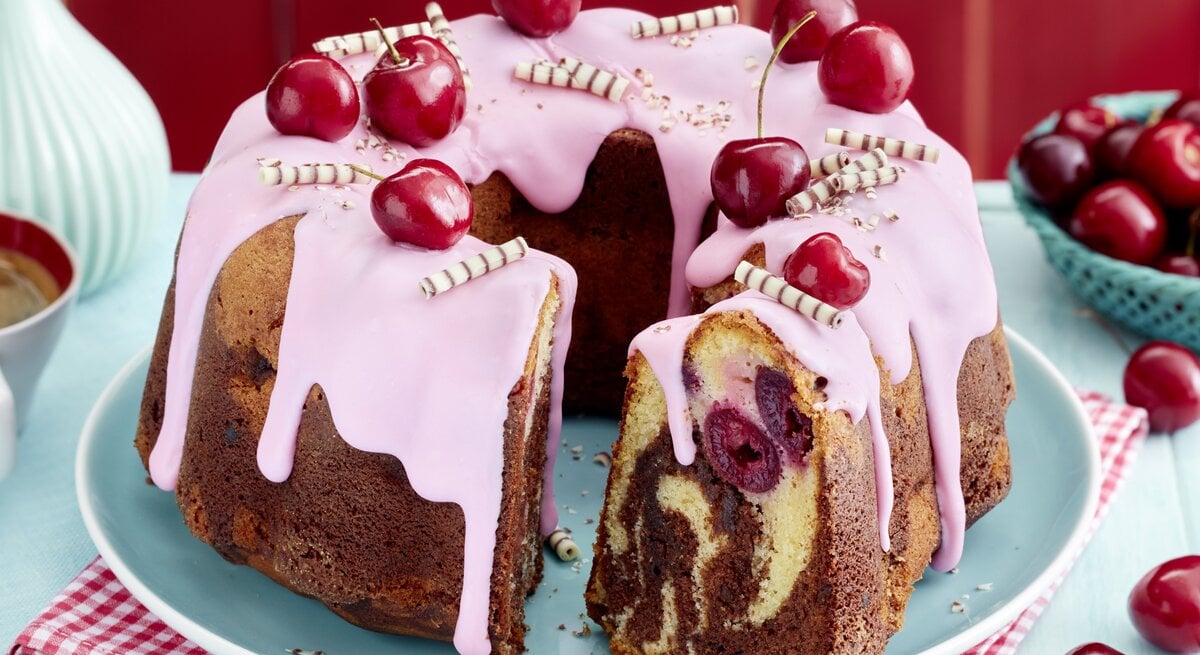 Ein saftiger Kirsch-Marmor-Hupf mit rosa Zuckerguss, frischen Kirschen und Schokoröllchen dekoriert, auf einem hellblauen Teller und rotem Untersetzer serviert. Im Anschnitt sind die Marmorierung und eingebackene Kirschen deutlich zu sehen.
