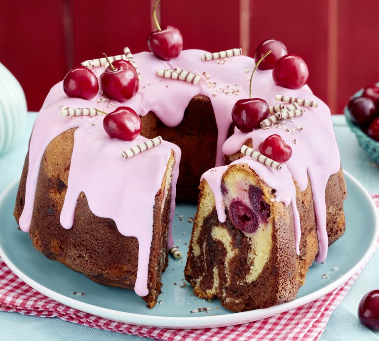 Ein saftiger Kirsch-Marmor-Hupf mit rosa Zuckerguss, frischen Kirschen und Schokoröllchen dekoriert, auf einem hellblauen Teller und rotem Untersetzer serviert. Im Anschnitt sind die Marmorierung und eingebackene Kirschen deutlich zu sehen. | © EAT CLUB/Isabell Triemer
