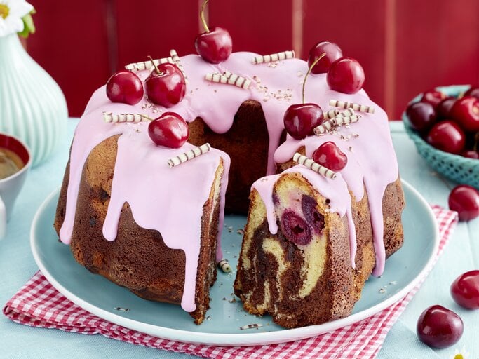 Ein saftiger Kirsch-Marmor-Hupf mit rosa Zuckerguss, frischen Kirschen und Schokoröllchen dekoriert, auf einem hellblauen Teller und rotem Untersetzer serviert. Im Anschnitt sind die Marmorierung und eingebackene Kirschen deutlich zu sehen. | © EAT CLUB/Isabell Triemer