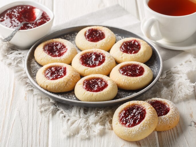 Frisch gebackene Marmeladen-Kekse mit Zuckerschnee serviert auf Leinentuch, daneben dampfender Tee in weißer Tasse. | © Getty Images / ALLEKO