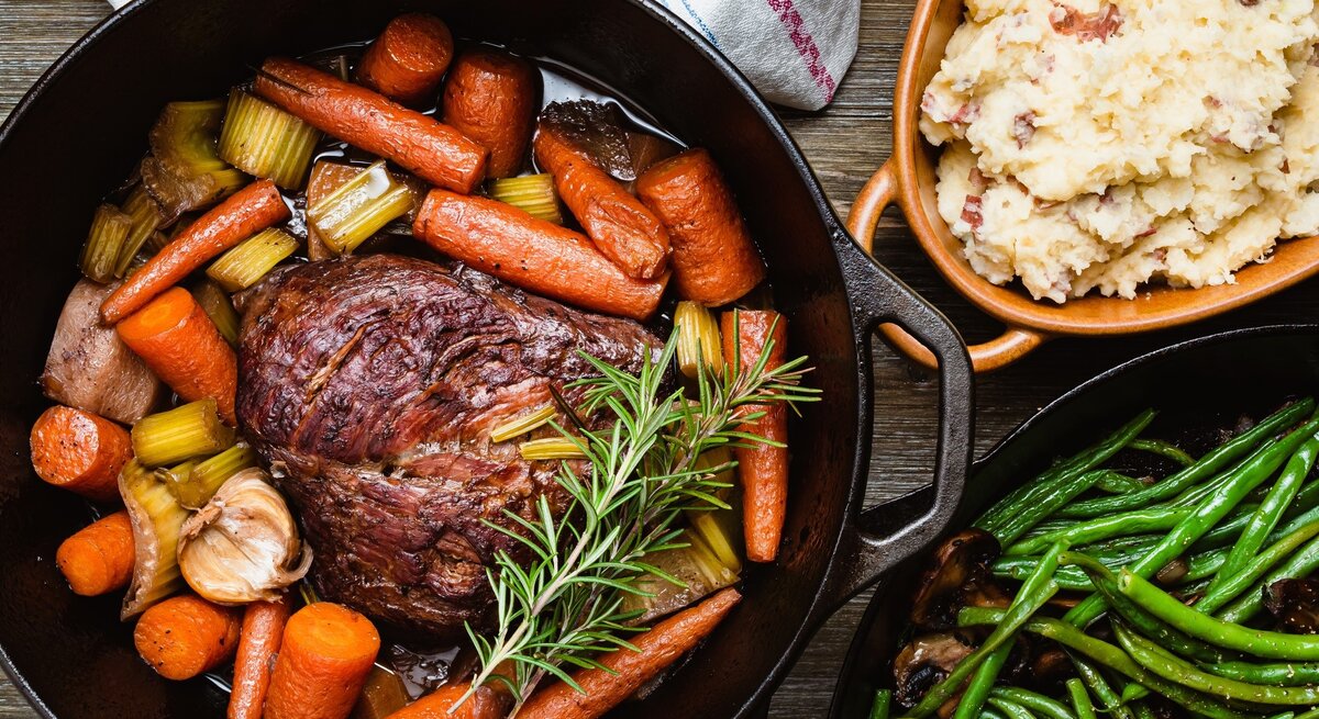 Brasato al Barolo, ein Schmorbraten aus Rindfleisch, in einem in einem gusseisernen Topf mit Griffen / Drumherum und ebenfalls im Topf Gemüse und Kräuter