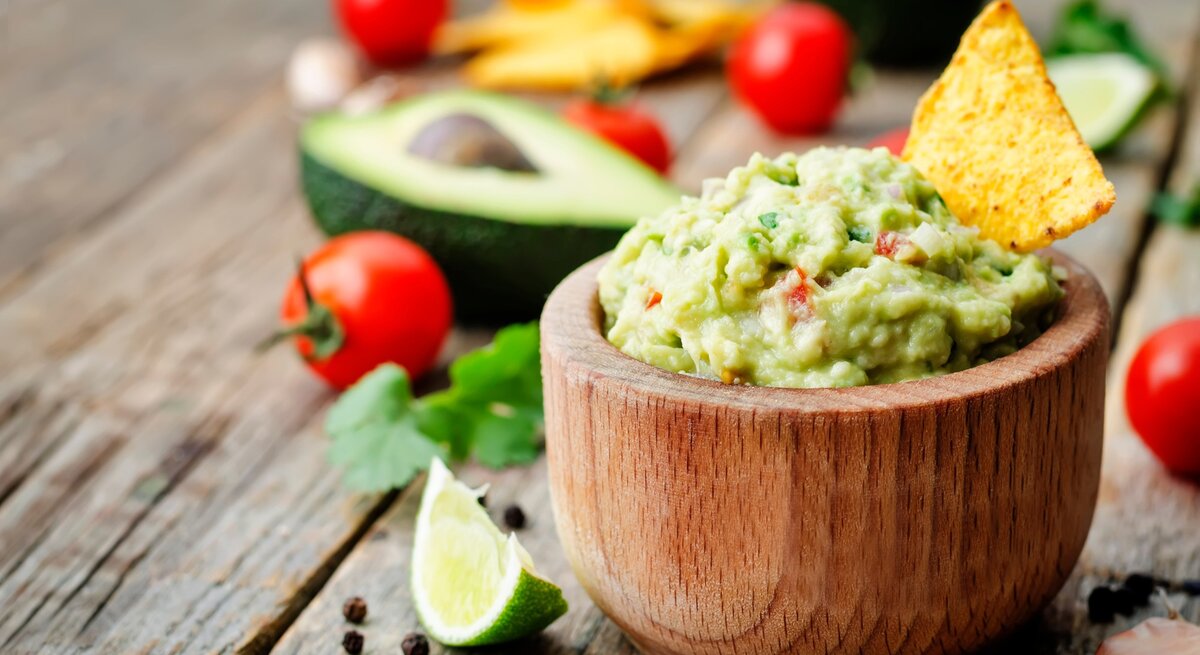 Guacamole in runder Holzschüssel / Nacho-Chip in der Guacamole / einzelne Tomaten, halbierte Avocado / Korianderblatt / Pfefferkörner / Untergrund grobes Holz