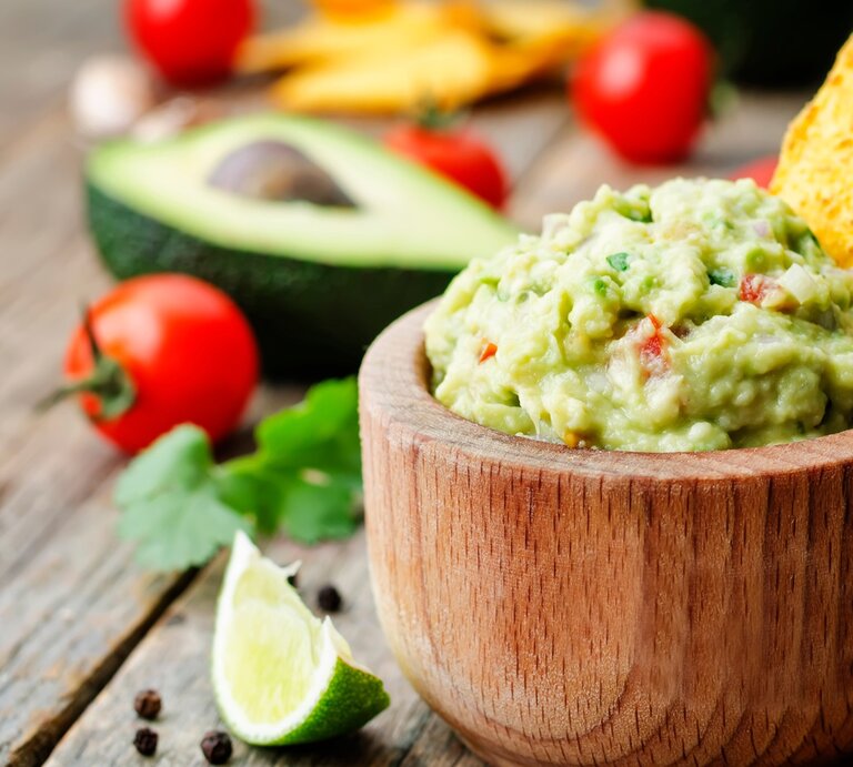 Guacamole in runder Holzschüssel / Nacho-Chip in der Guacamole / einzelne Tomaten, halbierte Avocado / Korianderblatt / Pfefferkörner / Untergrund grobes Holz | © Adobe Stock / nata_vkusidey