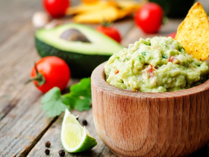 Guacamole in runder Holzschüssel / Nacho-Chip in der Guacamole / einzelne Tomaten, halbierte Avocado / Korianderblatt / Pfefferkörner / Untergrund grobes Holz | © Adobe Stock / nata_vkusidey