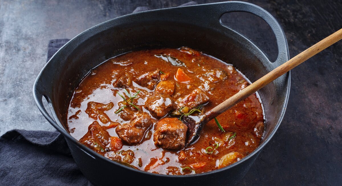 Ungarisches Gulasch wie bei Oma / Fleischstücke, Rosmarin, Möhrenstück / Holzlöffel im Topf / Draufsicht, schräg