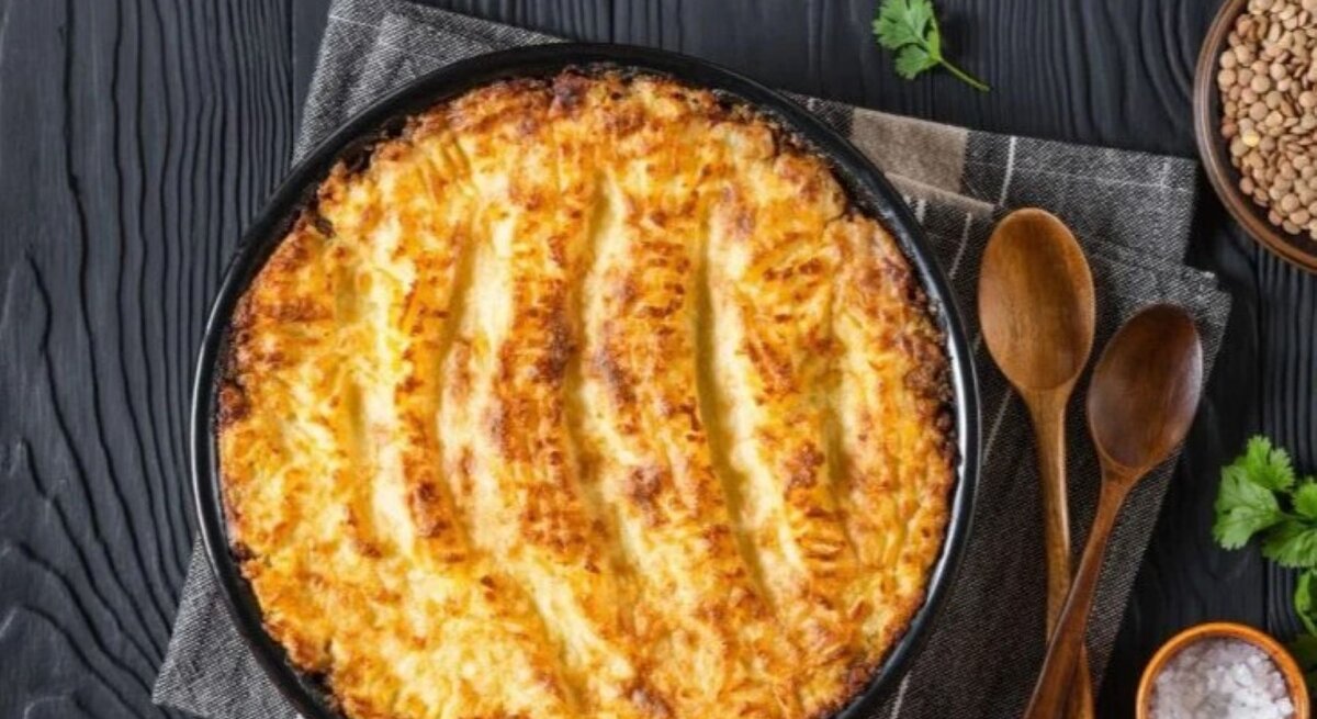 Veganer Linsen-Gemüse-Auflauf in runder Auflaufform auf dunklem Holztisch mit Linsen und Kräutern.