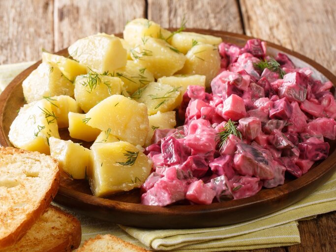 Roter Heringssalat, gekochte Kartoffeln, Dill auf runden Teller / zwei Scheiben Brot / Küchentuch / Untergrung grobes Holz / Draufsicht, schräg | © Adobe Stock / FomaA