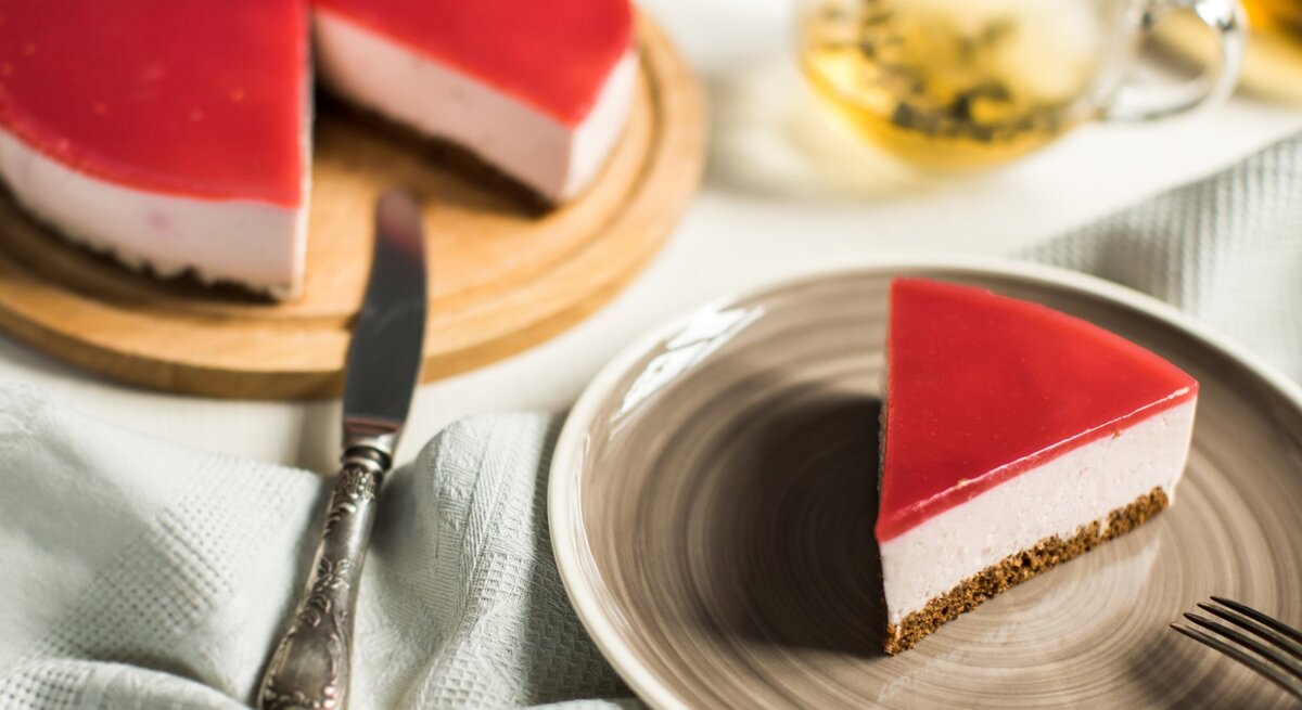 Ein Stück Schneewittchen-Torte auf einem braunen Teller. Links davon die Torte auf einem Tortenteller. Der Hintergrund ist hell. Links unten ein Geschirrtuch und rechts oben ein Tee-Glas. 