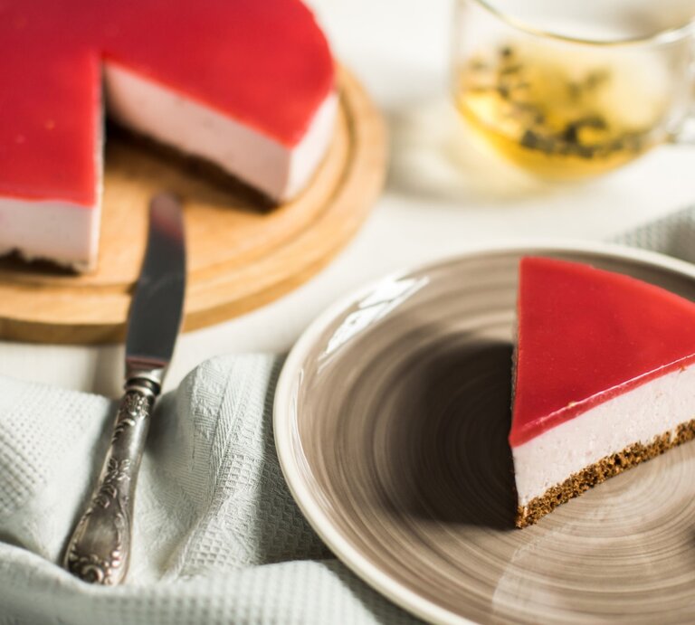 Ein Stück Schneewittchen-Torte auf einem braunen Teller. Links davon die Torte auf einem Tortenteller. Der Hintergrund ist hell. Links unten ein Geschirrtuch und rechts oben ein Tee-Glas.  | © GettyImages / yuliasverdlova