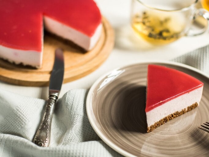 Ein Stück Schneewittchen-Torte auf einem braunen Teller. Links davon die Torte auf einem Tortenteller. Der Hintergrund ist hell. Links unten ein Geschirrtuch und rechts oben ein Tee-Glas.  | © GettyImages / yuliasverdlova