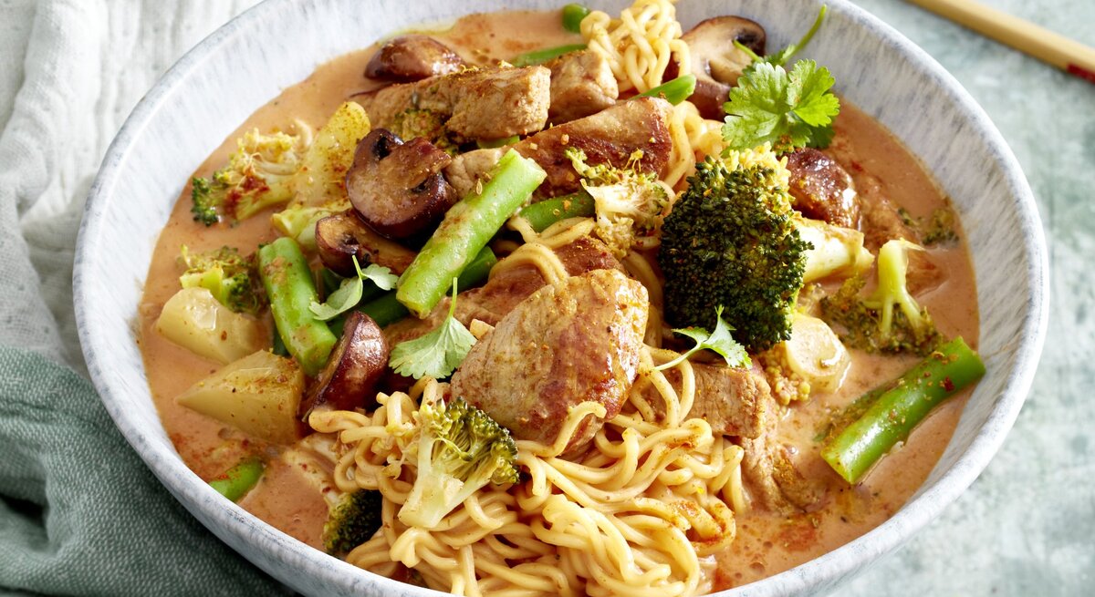 Thai-Curry mit Nudeln in einer hellgrauen Schüssel auf hellgrauem Hintergrund. Links ein Geschirrtuch, rechts Stäbchen. 