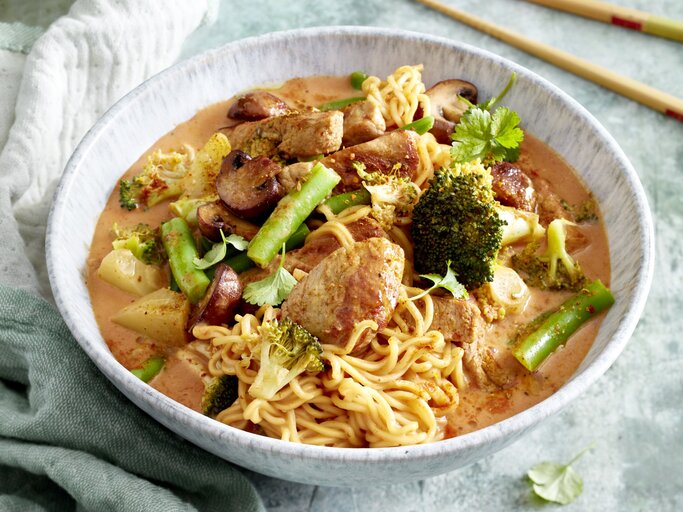 Thai-Curry mit Nudeln in einer hellgrauen Schüssel auf hellgrauem Hintergrund. Links ein Geschirrtuch, rechts Stäbchen.  | © Erlebe die Magie Thailands in einer Schüssel mit unseren aromatischen Thai-Curry-Nudeln - eine Reise für deine Geschmacksknospen, die du immer wieder erleben möchtest!