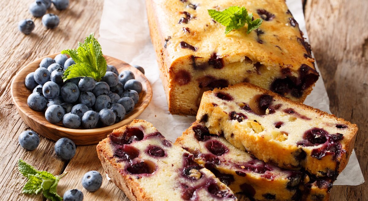 Zitronenkuchen mit Heidelbeeren / angeschnitten und einzelne Scheiben / runde Holzschale mit frischen Meidelbeeren / Minzblätter / Untergrund Holz / leicht schräge Drauf- und Frontalsicht