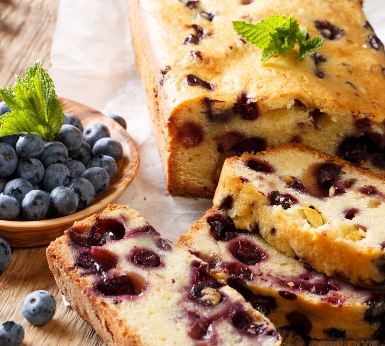 Zitronenkuchen mit Heidelbeeren / angeschnitten und einzelne Scheiben / runde Holzschale mit frischen Meidelbeeren / Minzblätter / Untergrund Holz / leicht schräge Drauf- und Frontalsicht | © Shutterstock / Sergii Koval