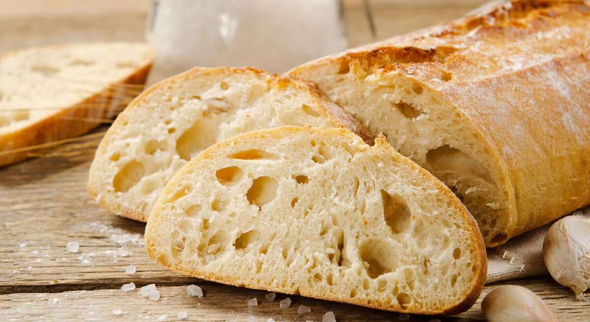 Ciabatta: zwei Scheinen und  der angeschnittene Brotlaib / einige Krümel Meersalz / Untergrund grobes Holt / Frontalsicht