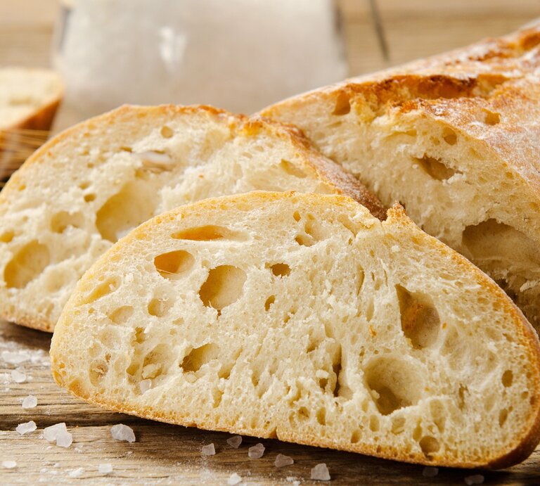 Ciabatta: zwei Scheinen und  der angeschnittene Brotlaib / einige Krümel Meersalz / Untergrund grobes Holt / Frontalsicht | © Adobe Stock / Mellisandra