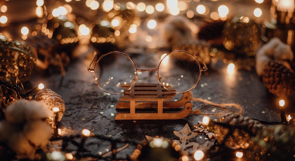 Runde goldene Brille von Harry Potter liegt auf einem kleinen Holzschlitten, umgeben von Tannenzapfen und warmen Lichterketten.