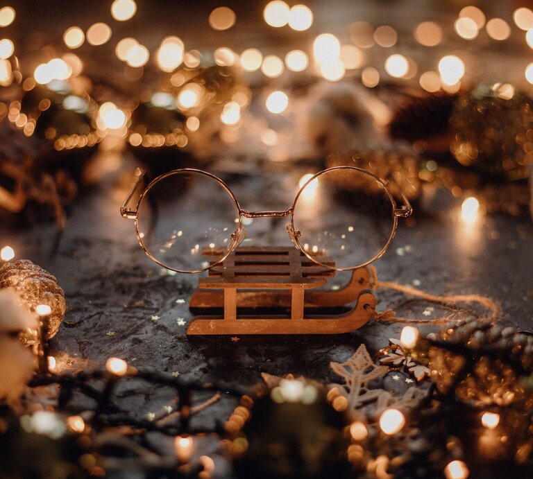 Runde goldene Brille von Harry Potter liegt auf einem kleinen Holzschlitten, umgeben von Tannenzapfen und warmen Lichterketten. | © AdobeStock/Barbara