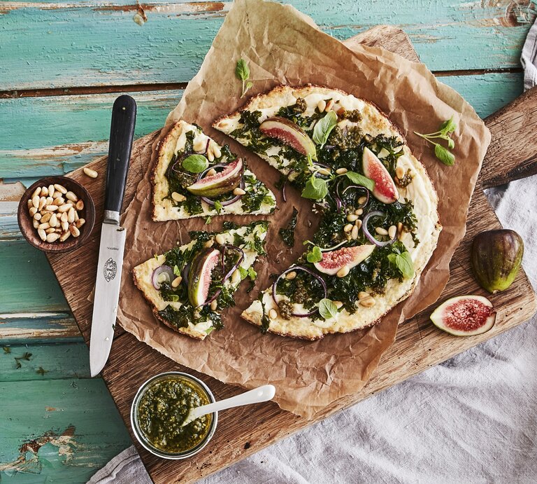 Draufsicht auf die Low-Carb-Pizza mit Grünkohlpest, Feigen und Pinienkernen, die auf einem Holzbrett auf einem türkisen Untergrund liegt. | © StockFood / Rådvik, Fanny
