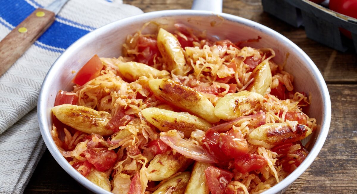 Schupfnudel-Sauerkraut-Pfanne auf hölzernem Hintergrund. Links Besteck und ein Geschirrtuch, oben auf dem Foto eine Tomate.