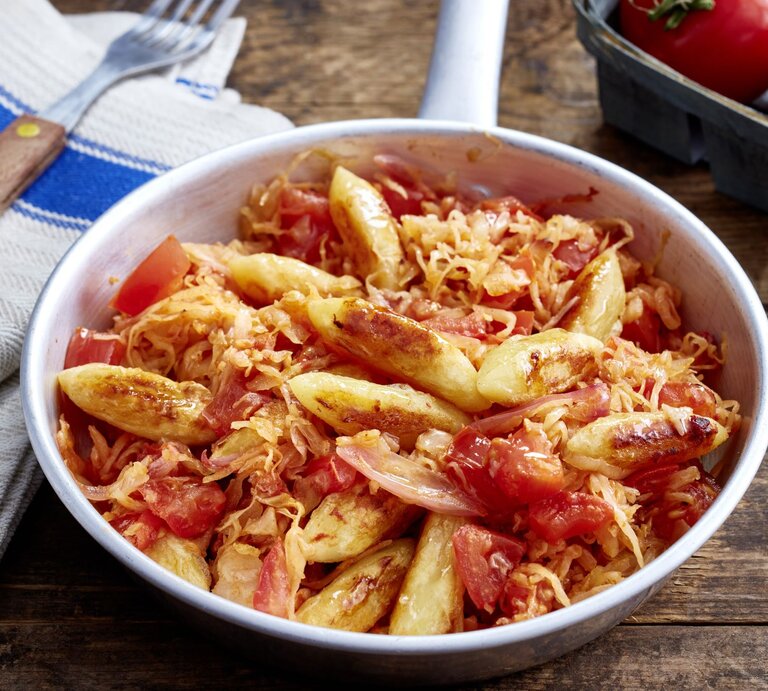 Schupfnudel-Sauerkraut-Pfanne auf hölzernem Hintergrund. Links Besteck und ein Geschirrtuch, oben auf dem Foto eine Tomate. | © EAT CLUB/Isabell Triemer