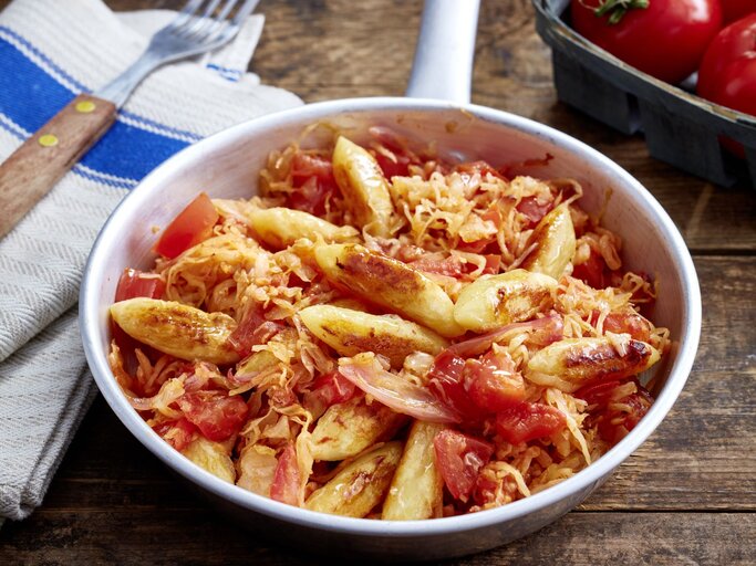 Schupfnudel-Sauerkraut-Pfanne auf hölzernem Hintergrund. Links Besteck und ein Geschirrtuch, oben auf dem Foto eine Tomate. | © EAT CLUB/Isabell Triemer
