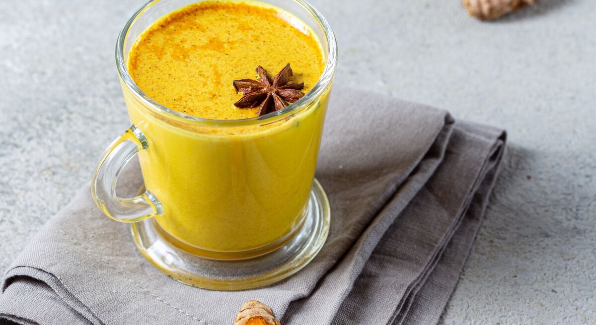 Ein Glas Goldene Milch, garniert mit einem Sternanis, steht auf einer grauen Stoffserviette. Daneben liegen frische Kurkuma- und Ingwerstücke.