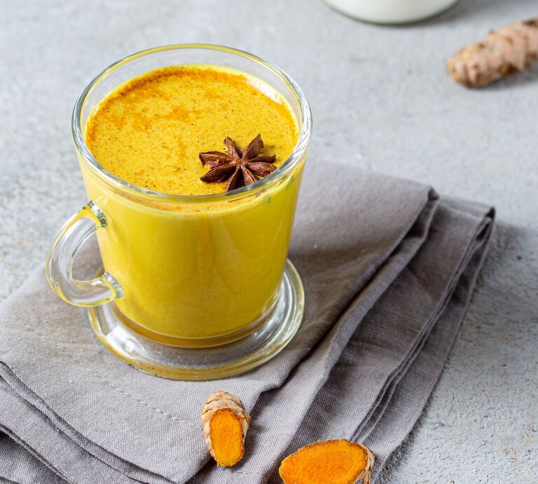 Ein Glas Goldene Milch, garniert mit einem Sternanis, steht auf einer grauen Stoffserviette. Daneben liegen frische Kurkuma- und Ingwerstücke. | © Getty Images/Larisa Blinova
