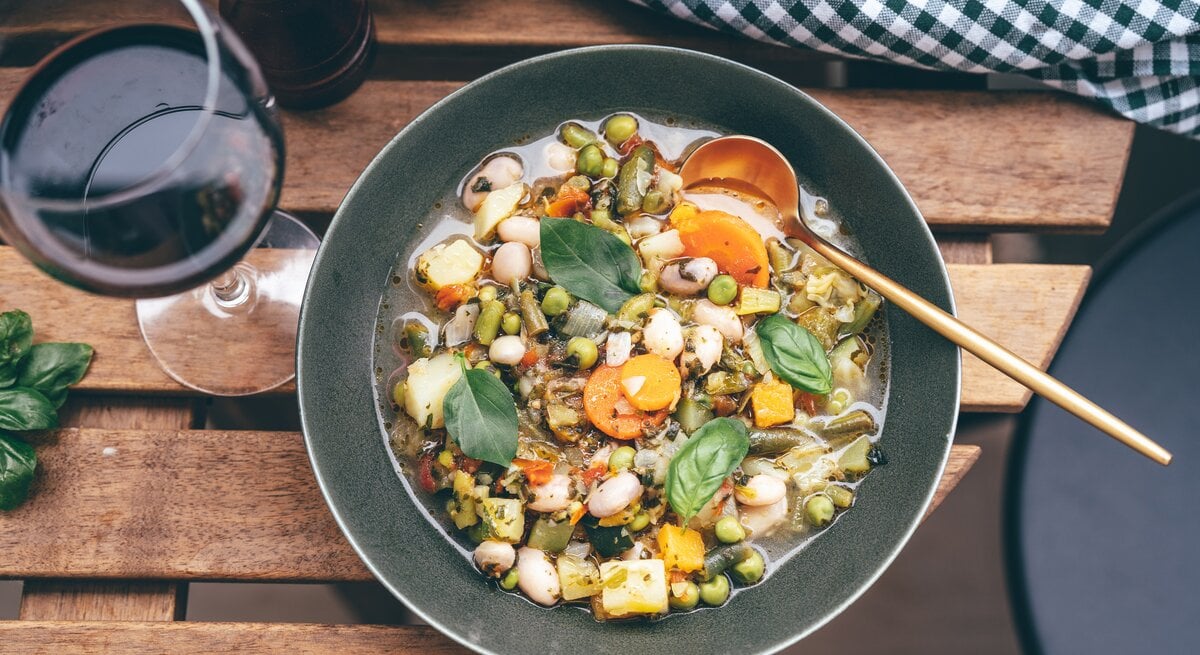Draufsicht auf einen tiefen, dunklen Teller mit einer Minestrone aus viel Gemüse, der auf einem Tisch neben einem Weinglas steht.