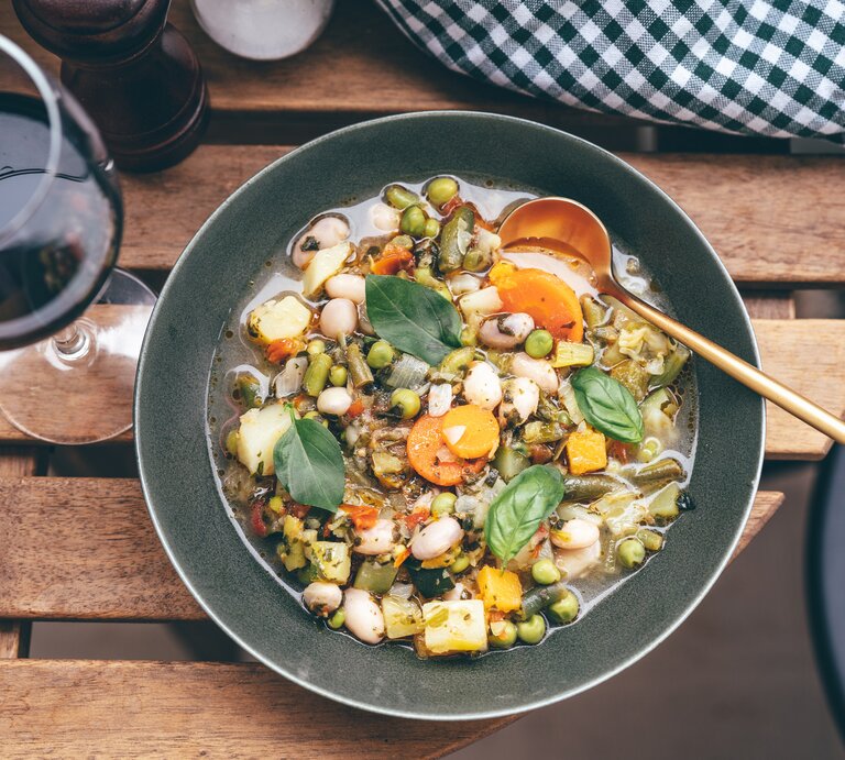 Draufsicht auf einen tiefen, dunklen Teller mit einer Minestrone aus viel Gemüse, der auf einem Tisch neben einem Weinglas steht. | © Getty Images / Yummy pic