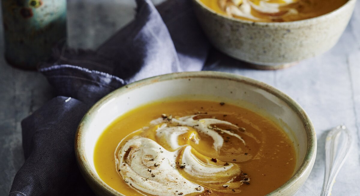 Zwei Schalen Kürbissuppe mit Sahne und Zimt stehen auf grauem Stein, daneben ein Löffel, Leinentuch und frisches Sauerteigbrot.