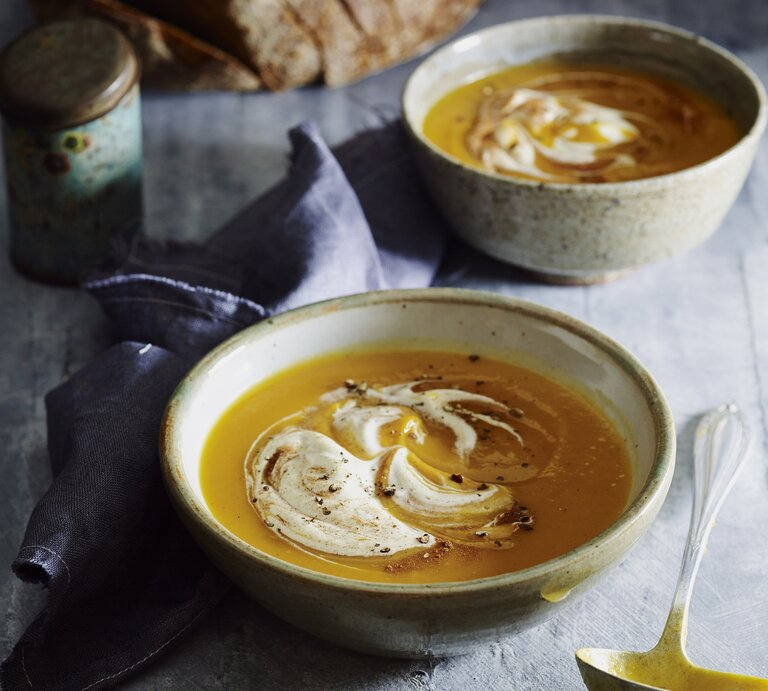 Zwei Schalen Kürbissuppe mit Sahne und Zimt stehen auf grauem Stein, daneben ein Löffel, Leinentuch und frisches Sauerteigbrot. | © StockFood / Are Media / Are Media
