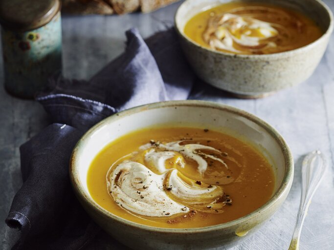 Zwei Schalen Kürbissuppe mit Sahne und Zimt stehen auf grauem Stein, daneben ein Löffel, Leinentuch und frisches Sauerteigbrot. | © StockFood / Are Media / Are Media