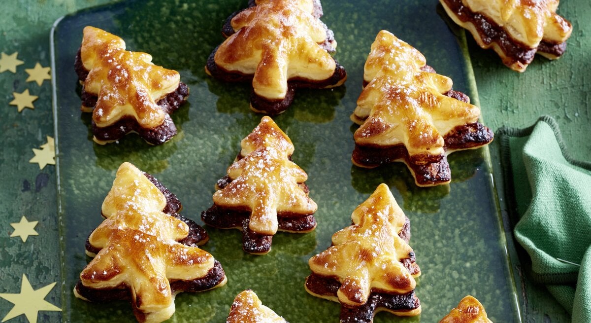 Schräge Draufsicht auf ein Blech mit frisch gebackenen Nougat-Tannenbäume vor einer weihnachtlichen Kulisse.